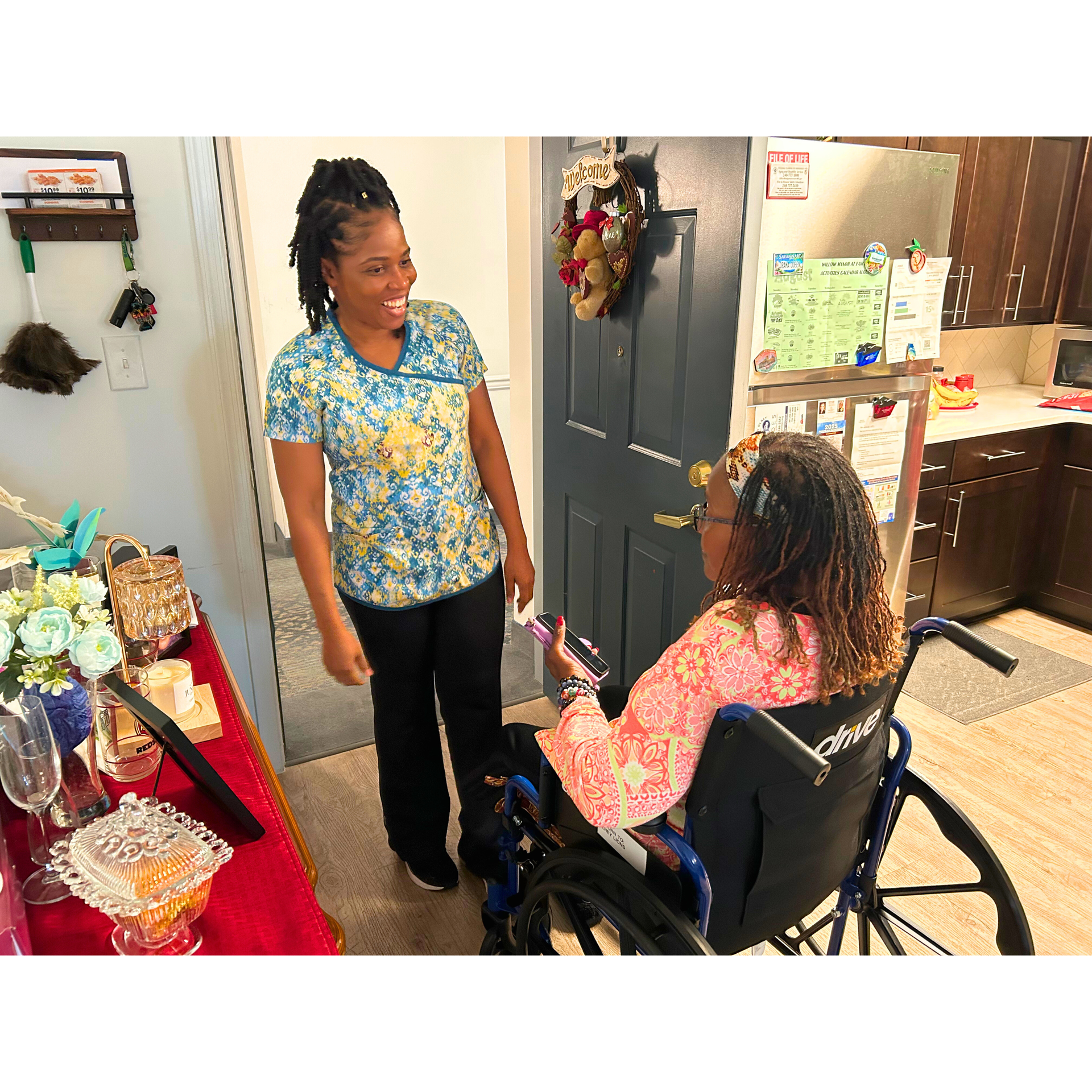 Woman in blue shirt speaking with a seated person in a wheelchair in a room with snacks and kitchen appliances