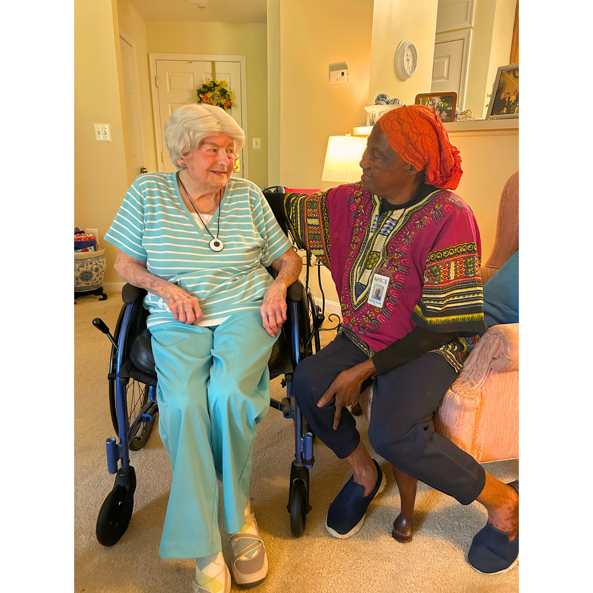 Two women in conversation indoors; one seated in a wheelchair, the other has her arm around her.