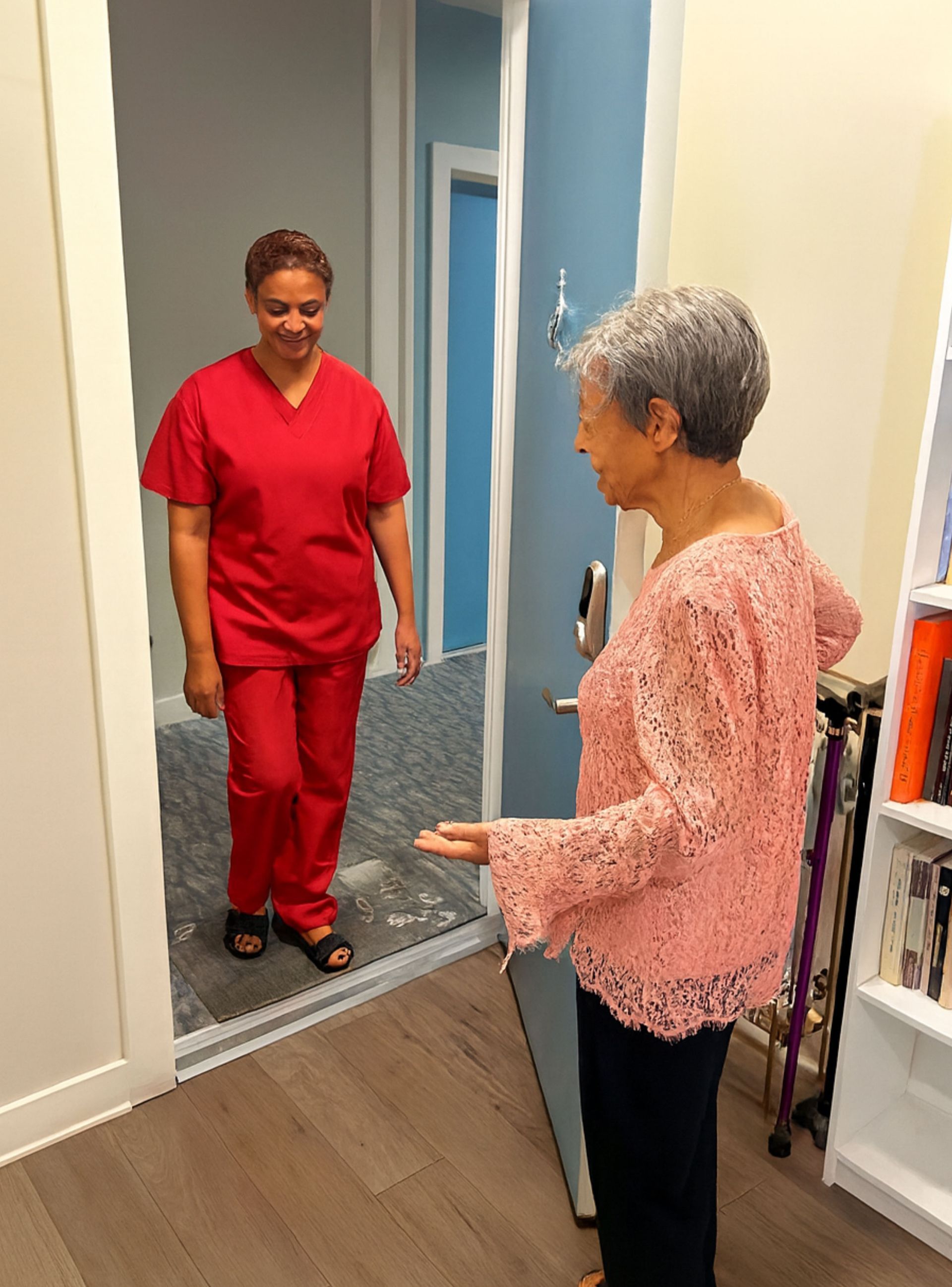 Woman in red scrubs enters a room where another woman gestures toward her. Interior setting with books.