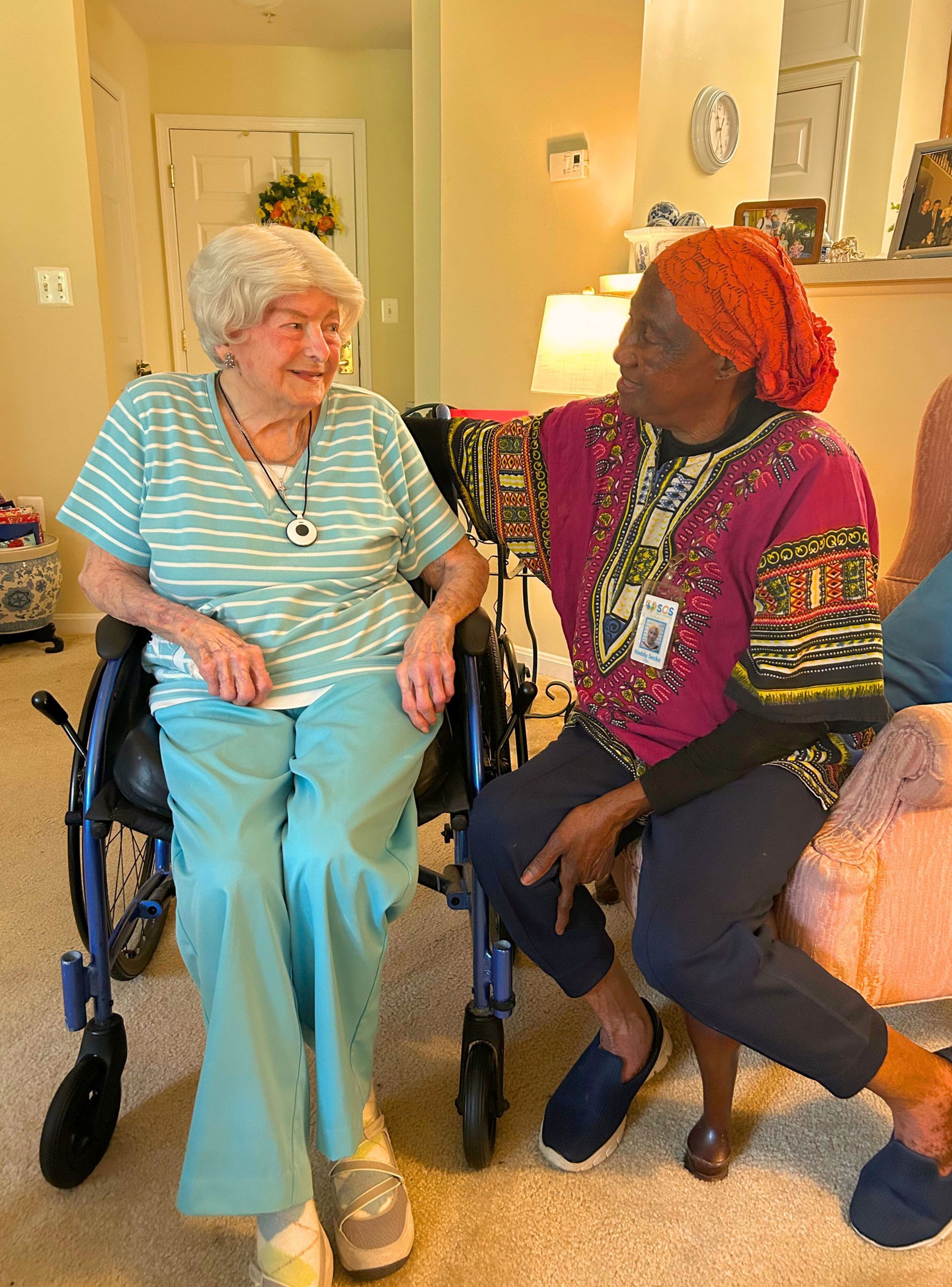 Two people sit and converse indoors. One is in a wheelchair wearing a blue and white striped shirt. The other wears an orange head covering and a colorful shirt.