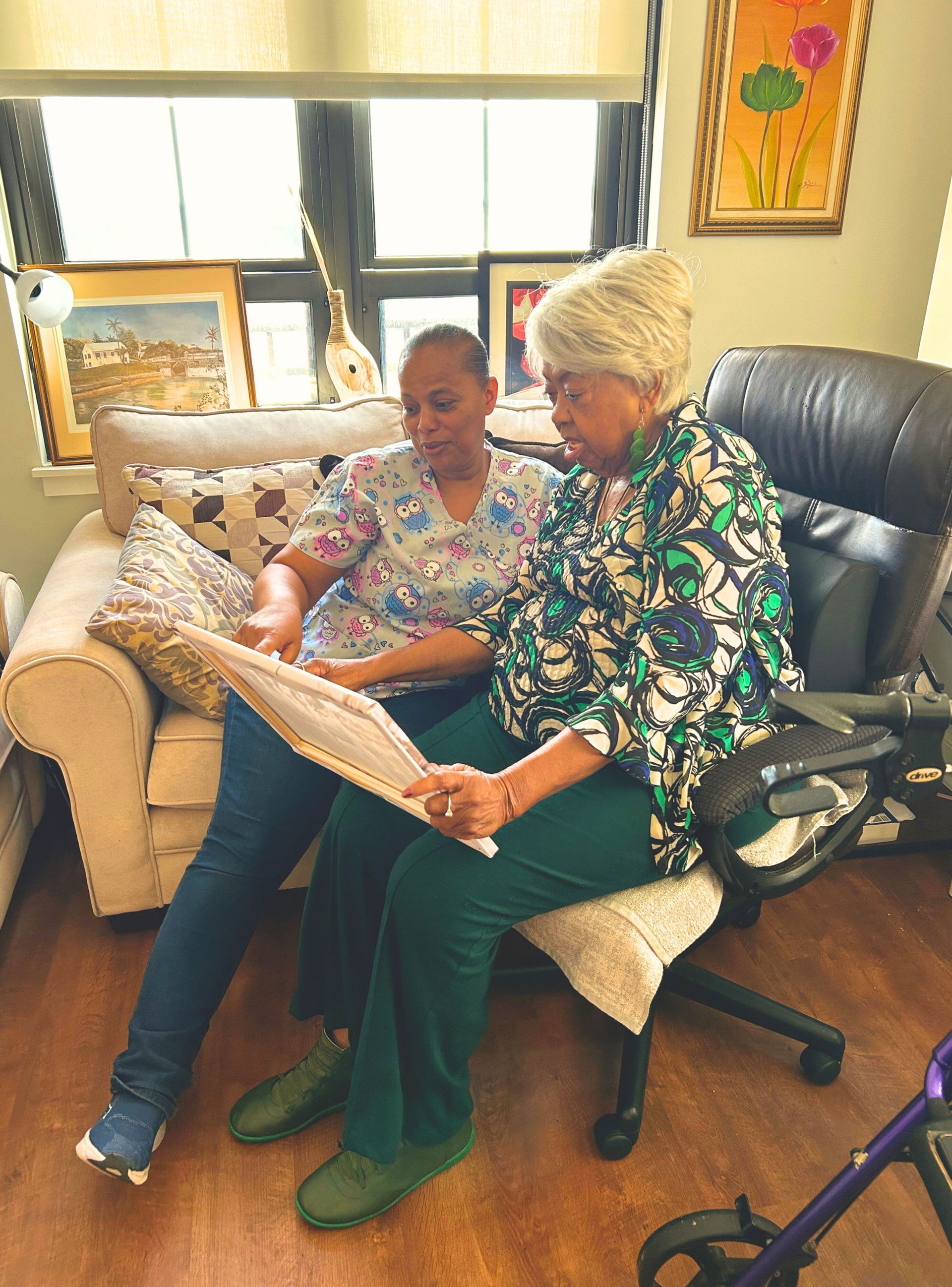 Two women looking at a framed picture. One is sitting in a chair; the other is beside her on a couch.