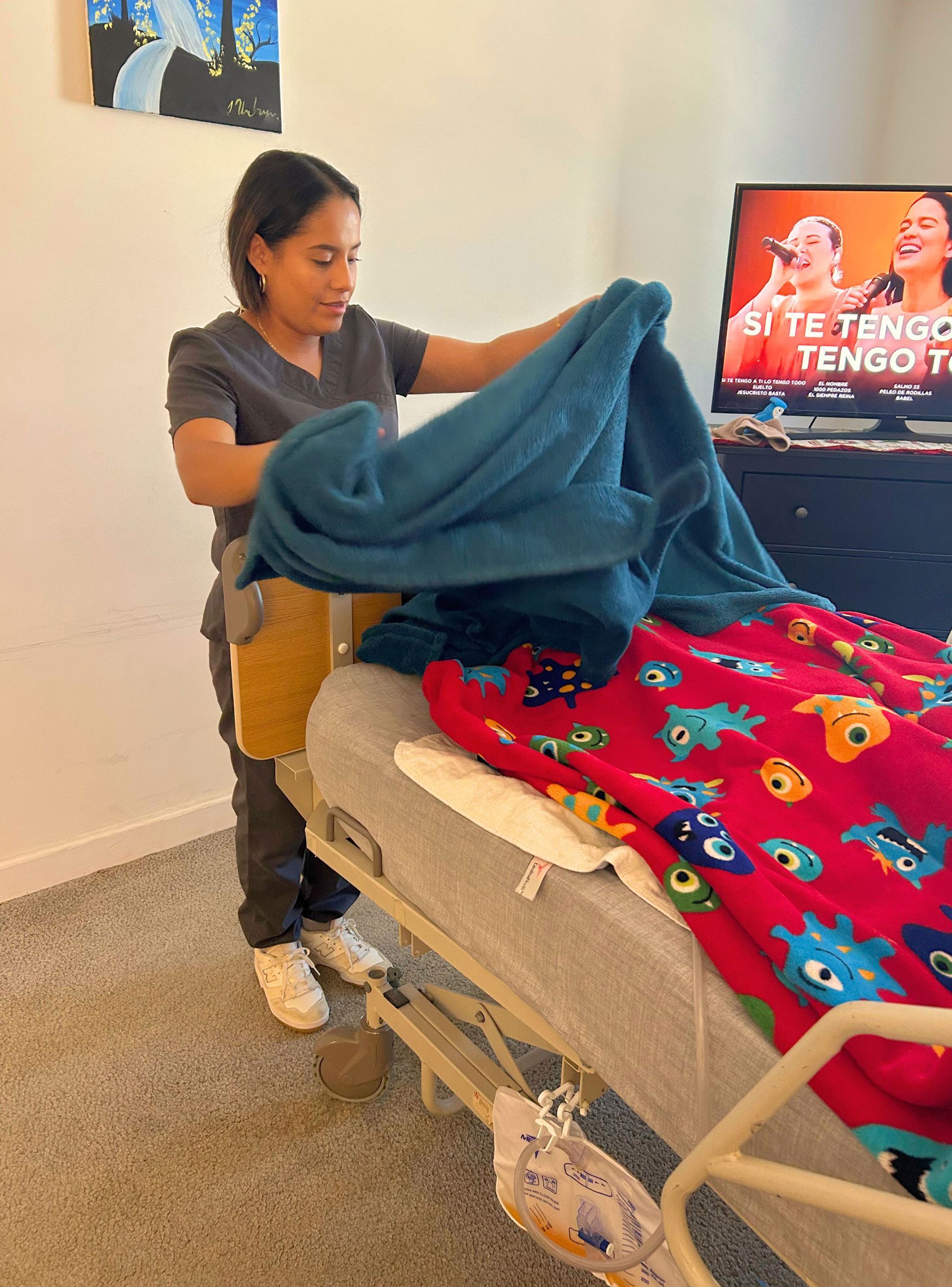 Person in scrubs folds a blue blanket over a bed with a red monster blanket, inside a home.