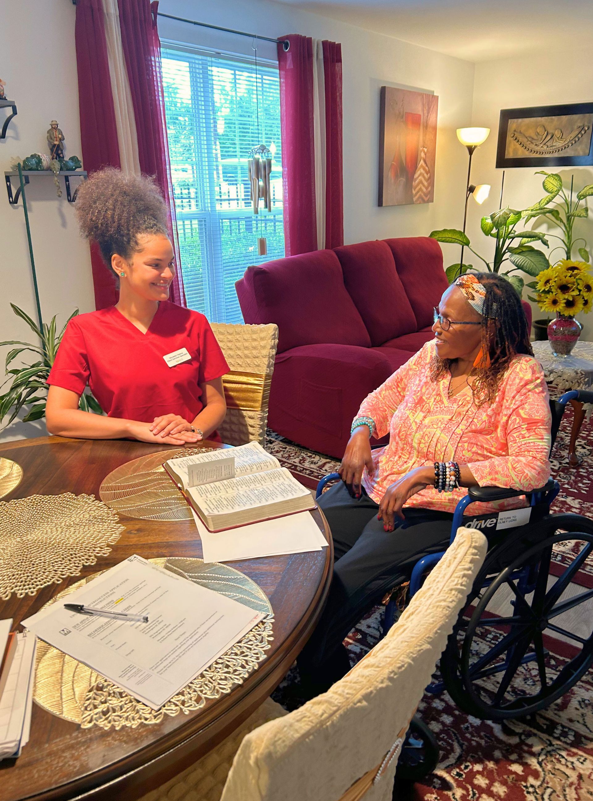 Woman in wheelchair sits with person in red shirt at a round table, discussing. Indoor living room setting.