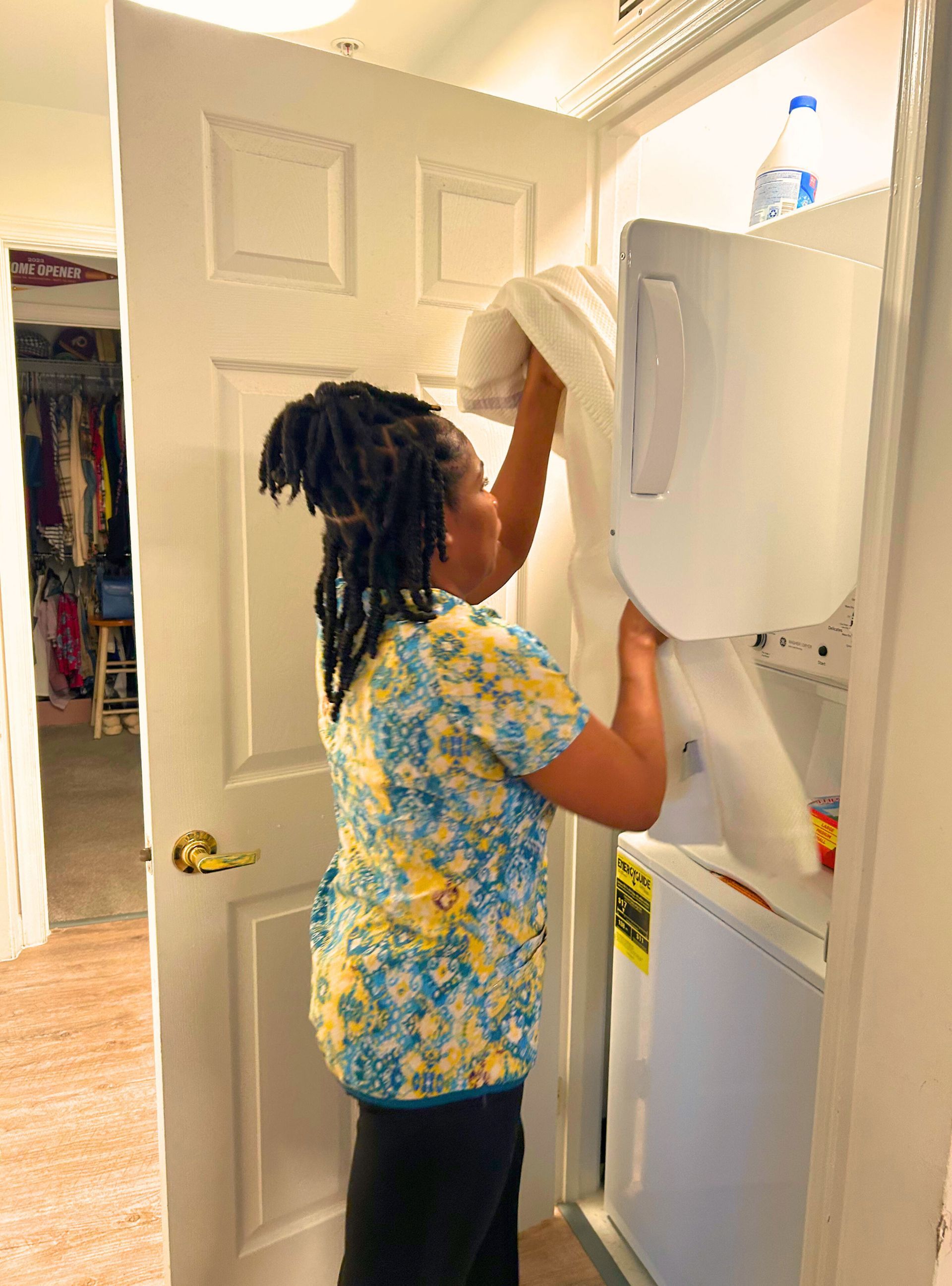 Woman putting a white garment into a large, open, upright freezer in a room with a closed door on the left.
