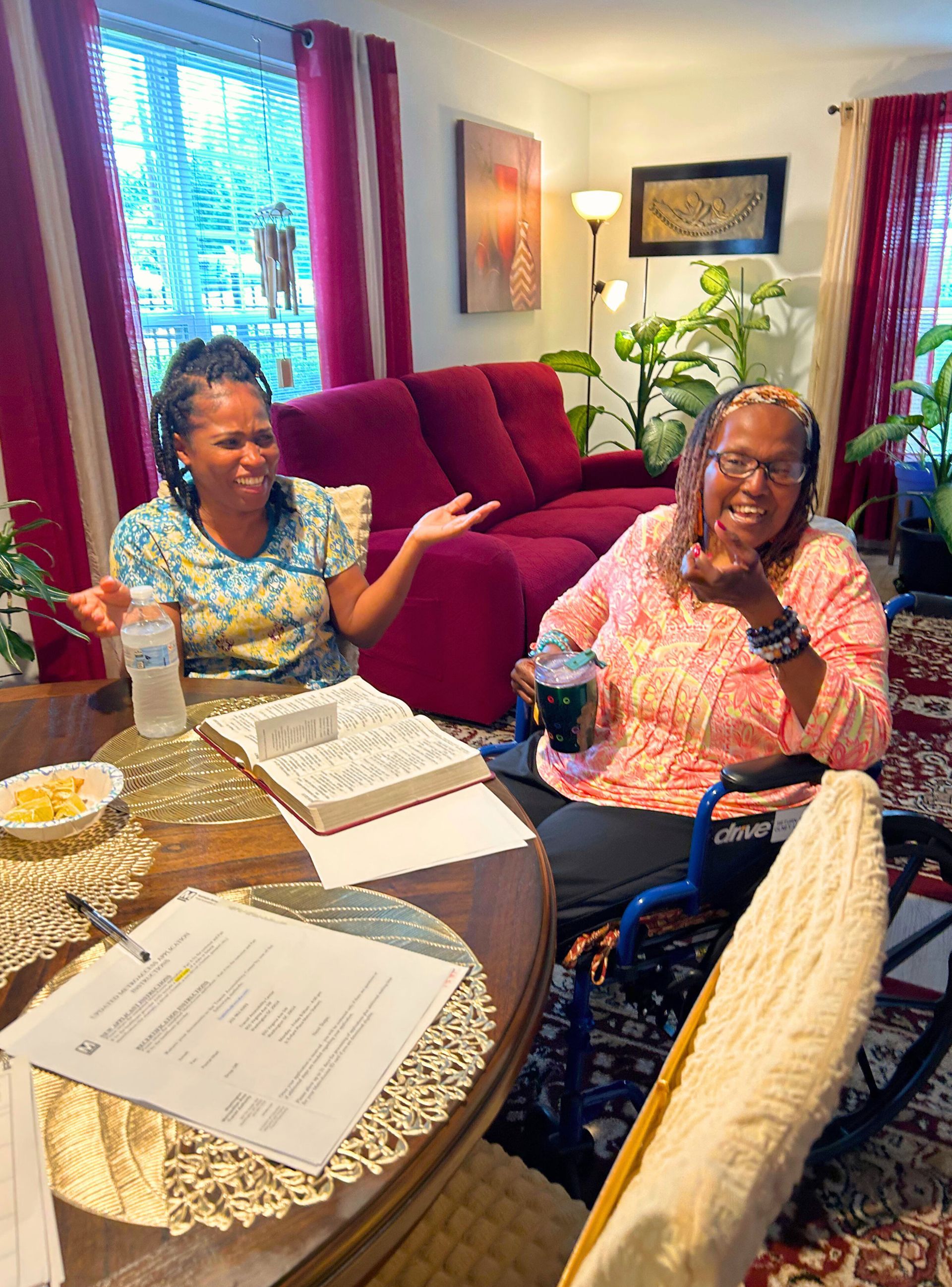 Two women at table. One in wheelchair, holding drink; the other gesturing. Red couch and curtains.
