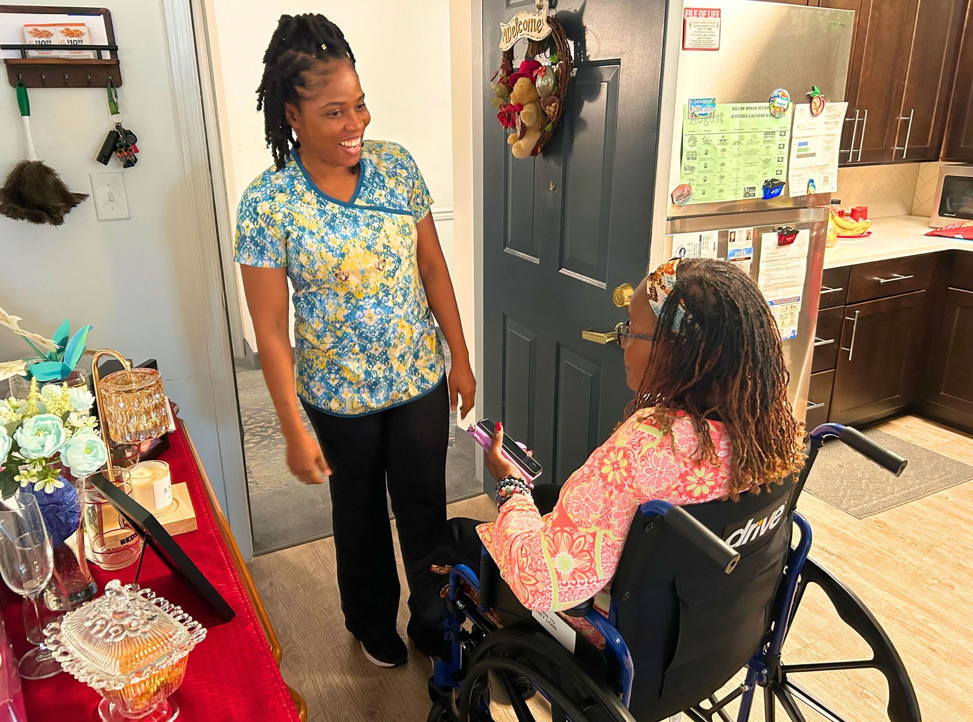 Woman in scrubs smiles at another in a wheelchair; they are indoors.
