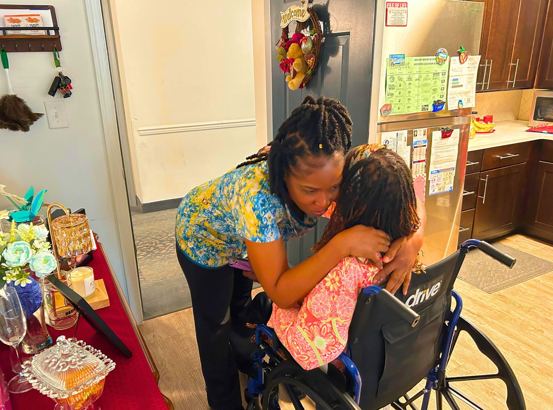 Woman hugging a child in a wheelchair inside a home kitchen. The woman leans in to embrace.