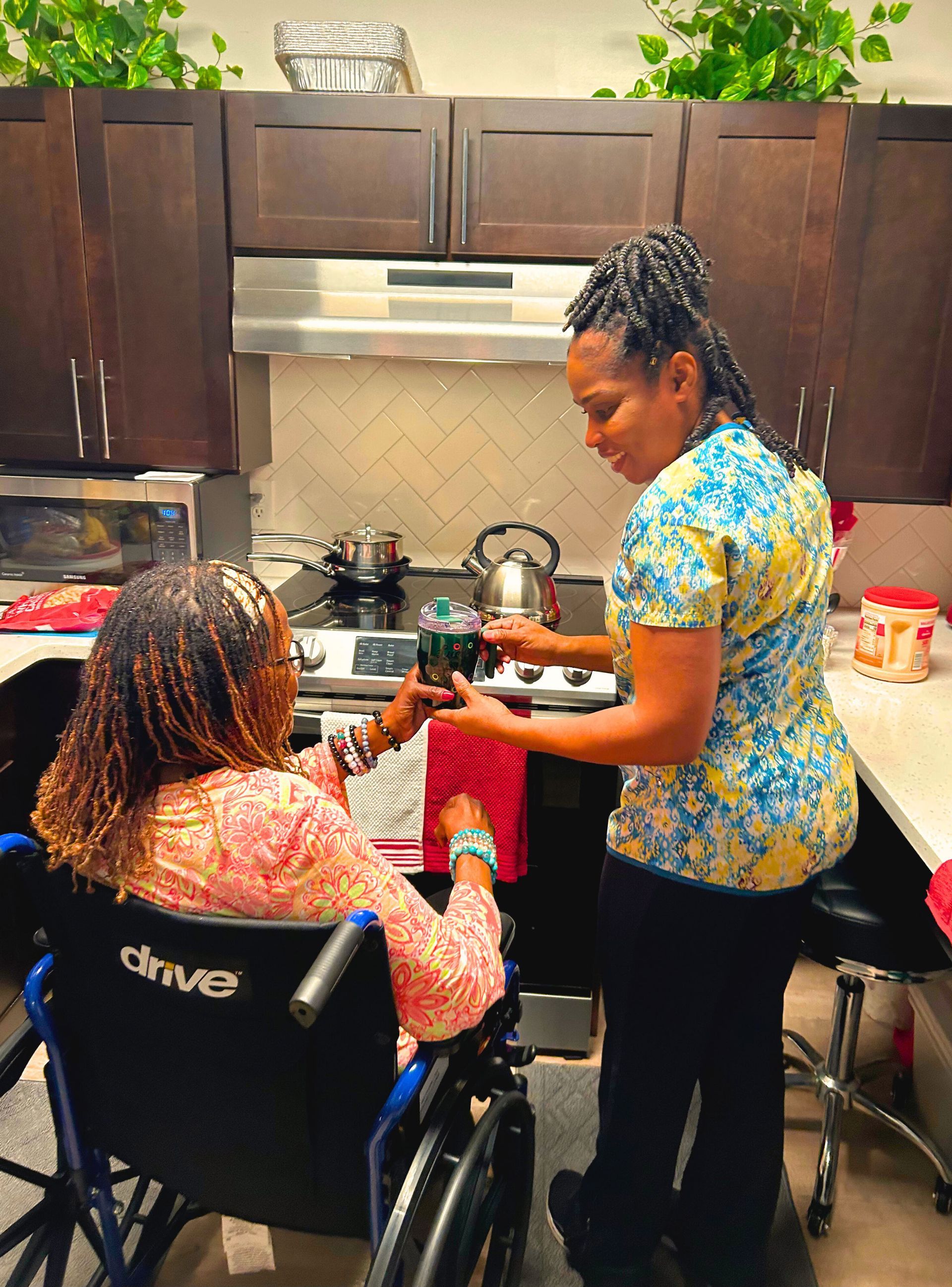 Woman in wheelchair, kitchen. Another woman hands her a drink. Cabinets, stove, and plants are visible.