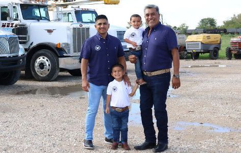 Four people pose near trucks: two men, and two young children, smiling, in front of a business.