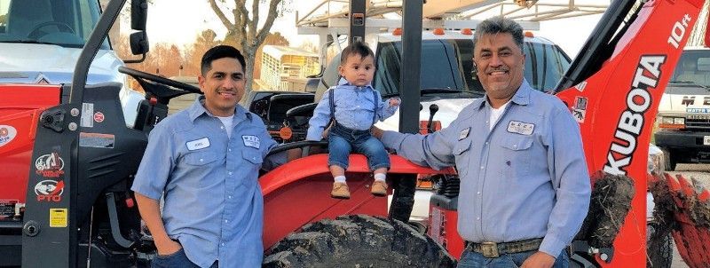 Two men and a baby on a red tractor. Men in blue shirts, baby in blue shirt, suspenders. Smiles. Outdoor setting.
