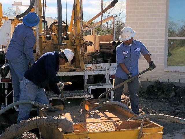 Three workers operating well drilling equipment near a building.
