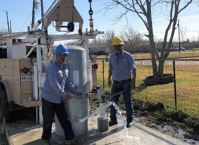 Two men in work clothes operate a well, water flowing. One pumps, the other observes. Outdoors.