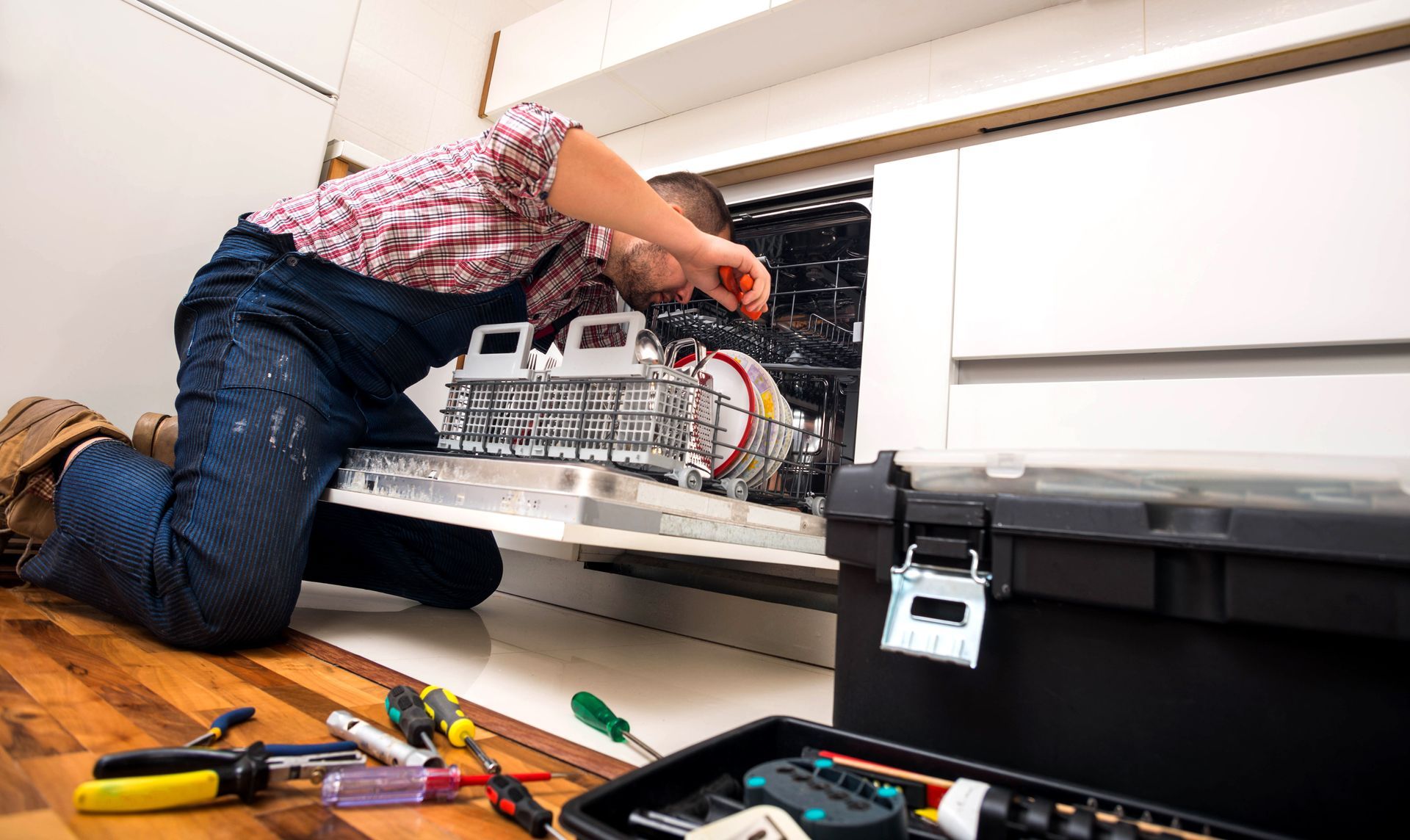 A person in overalls repairs a dishwasher, with a toolbox and tools on the floor in a kitchen.