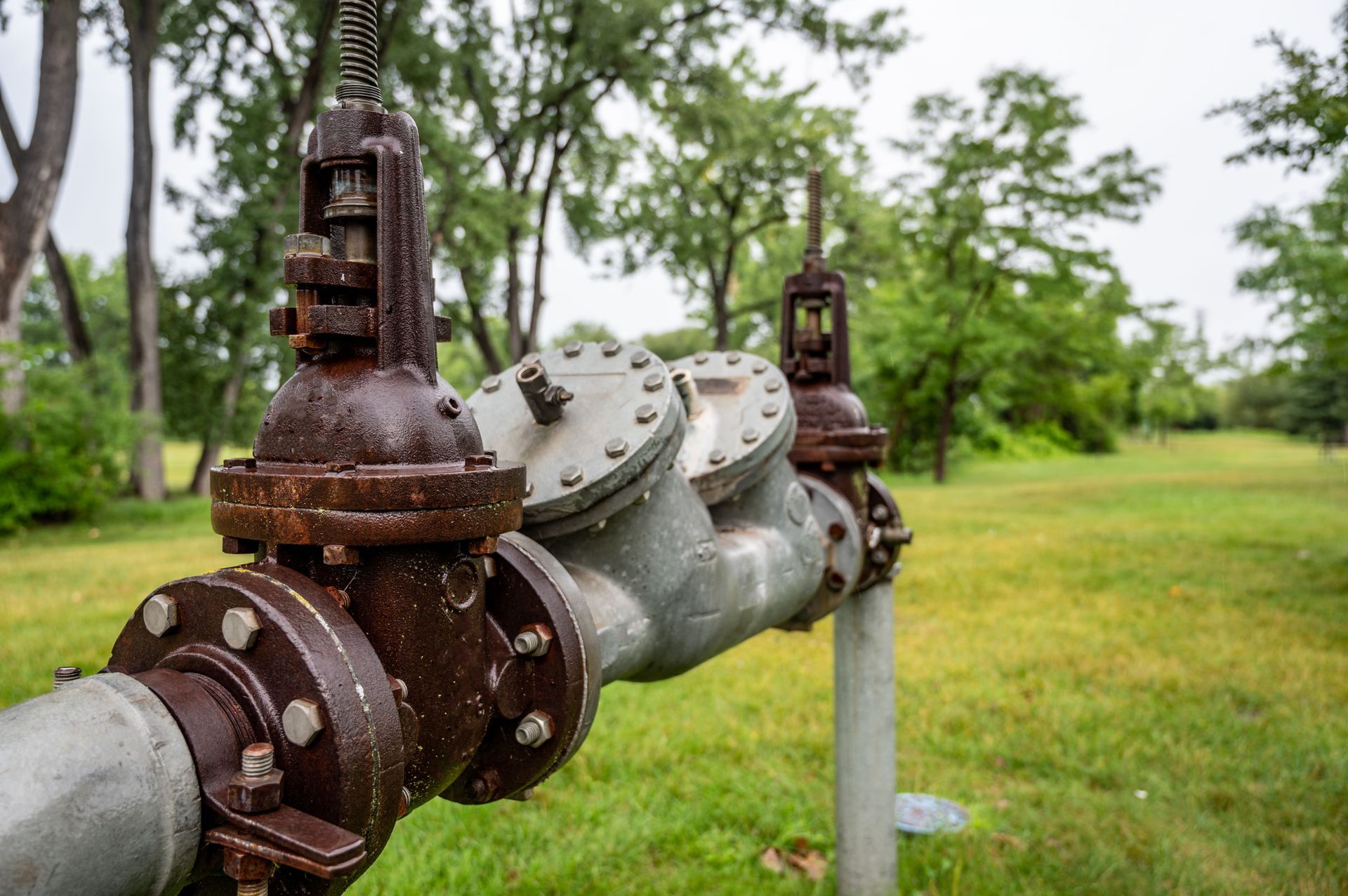 Rusty pipes with valves in a grassy field, trees in the background.