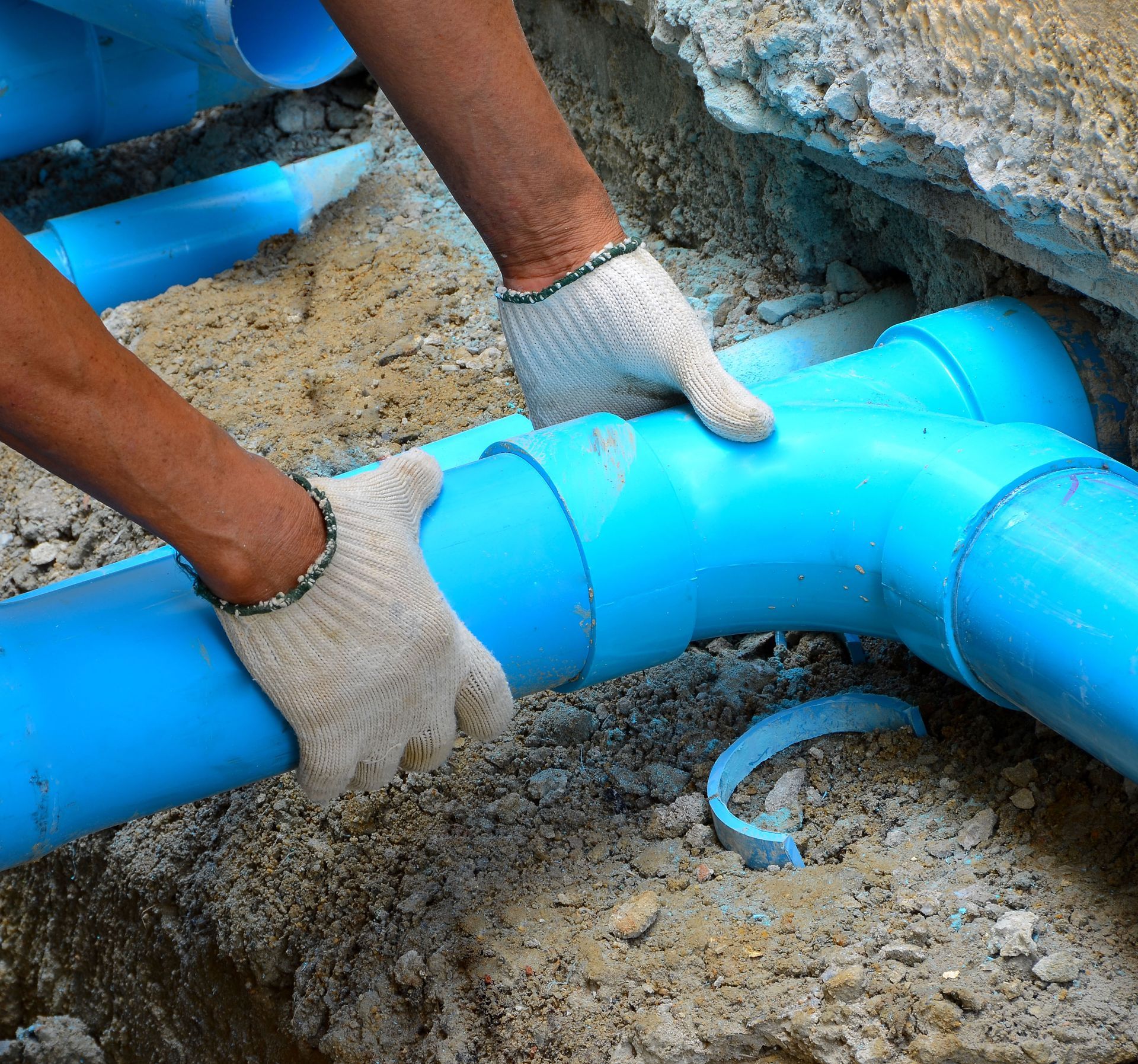 Hands wearing gloves connect blue PVC pipes in a trench.