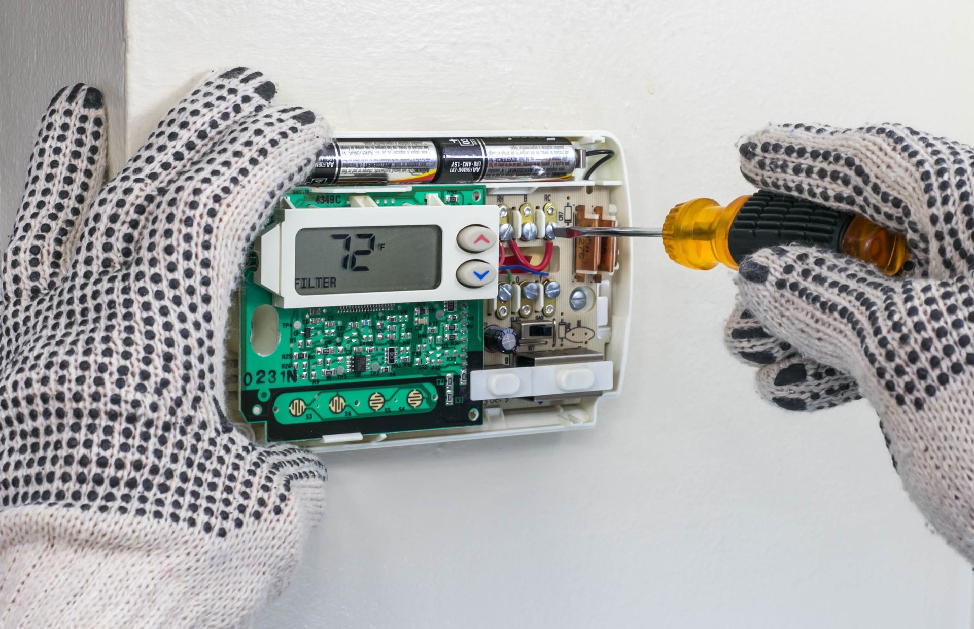 Hands wearing gloves, using a screwdriver to work on a thermostat mounted on a white wall.