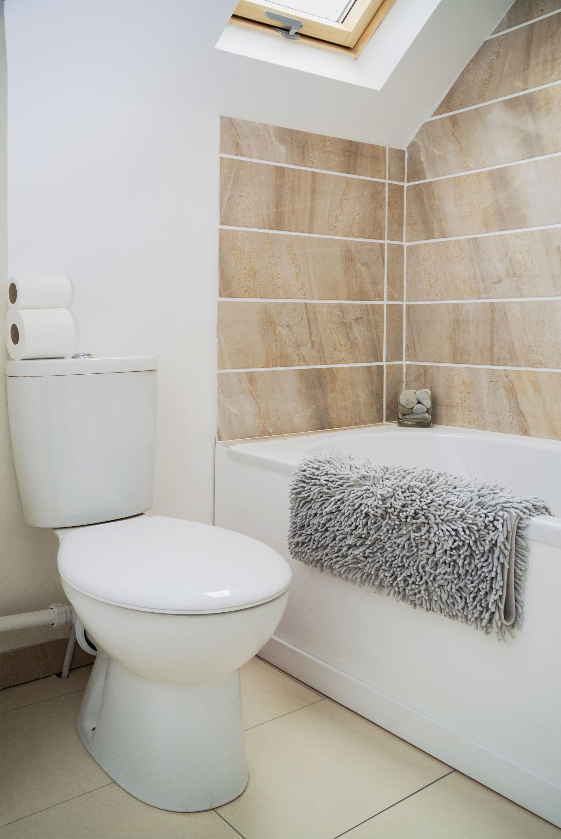 Toilet in a bathroom with tan tiled walls and a bathtub with a gray rug.