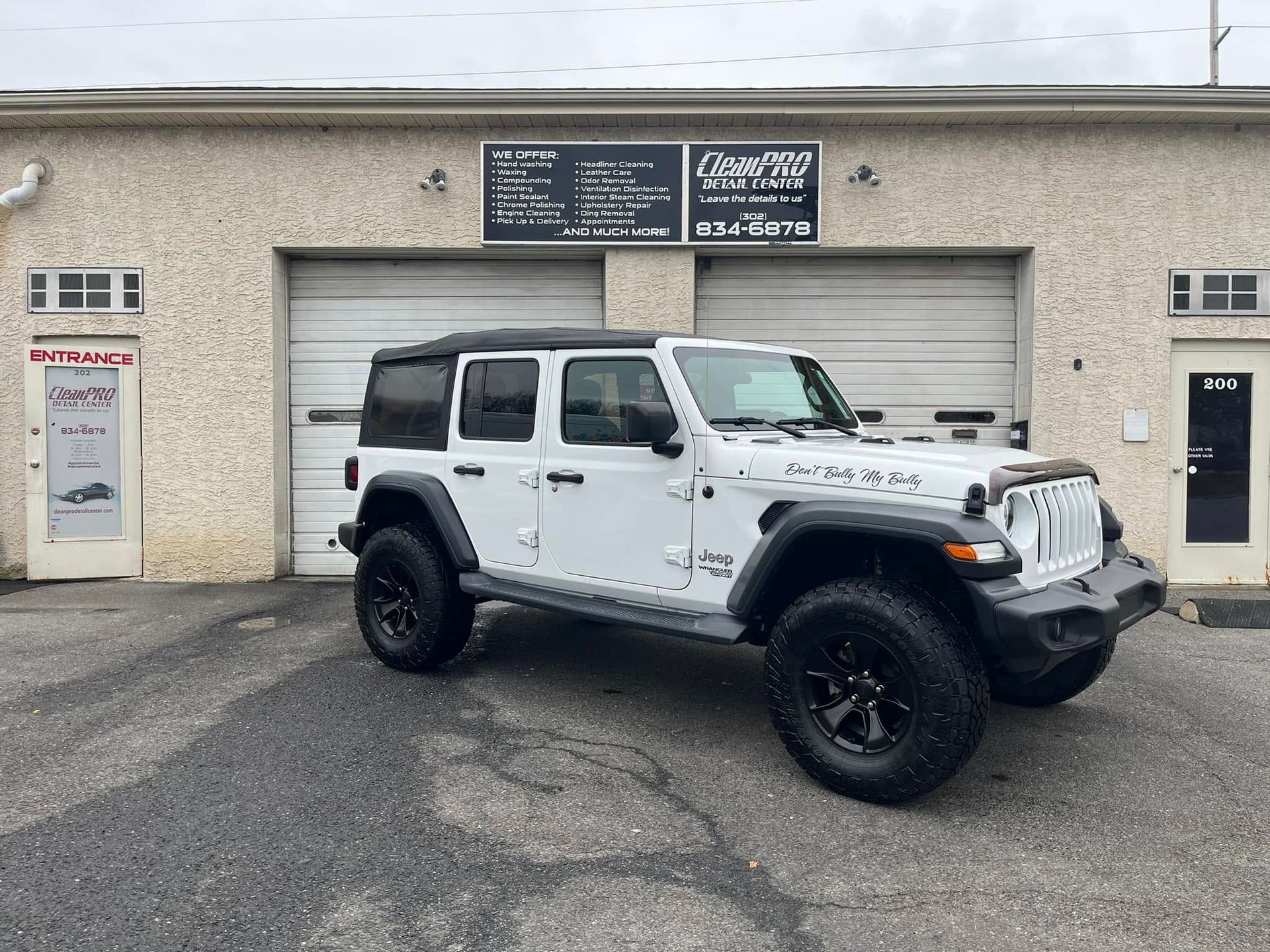 White four-door Jeep Wrangler parked in front of a beige auto shop with garage doors.