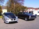 Two cars parked on a driveway, a silver sedan and a black classic car, with houses in the background