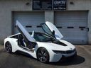 White sports car with butterfly doors open, parked in front of a garage.
