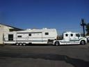 White pickup truck towing a large white camper trailer on a roadside under a blue sky