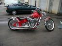 Red chopper-style motorcycle with chrome accents parked on a street beside a dark green car.