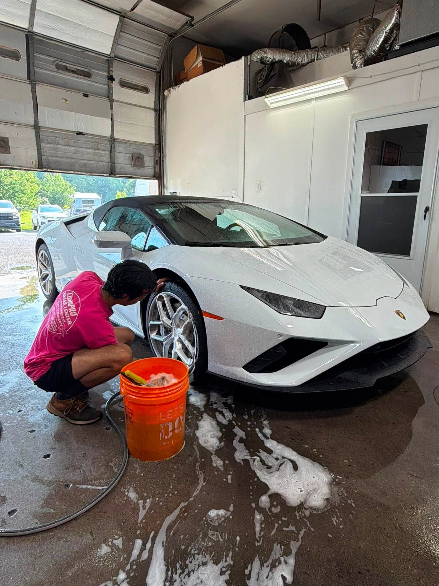 Person washing a white sports car in a garage with an orange bucket and soap suds