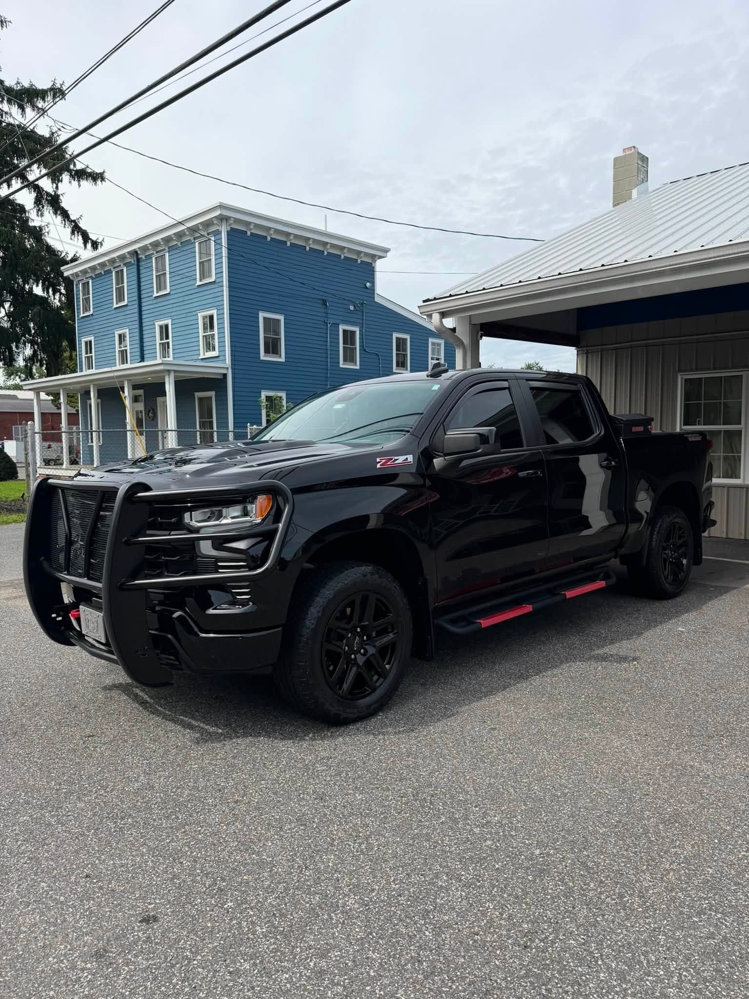 Black pickup truck with off-road lights parked on a street beside a blue building and house