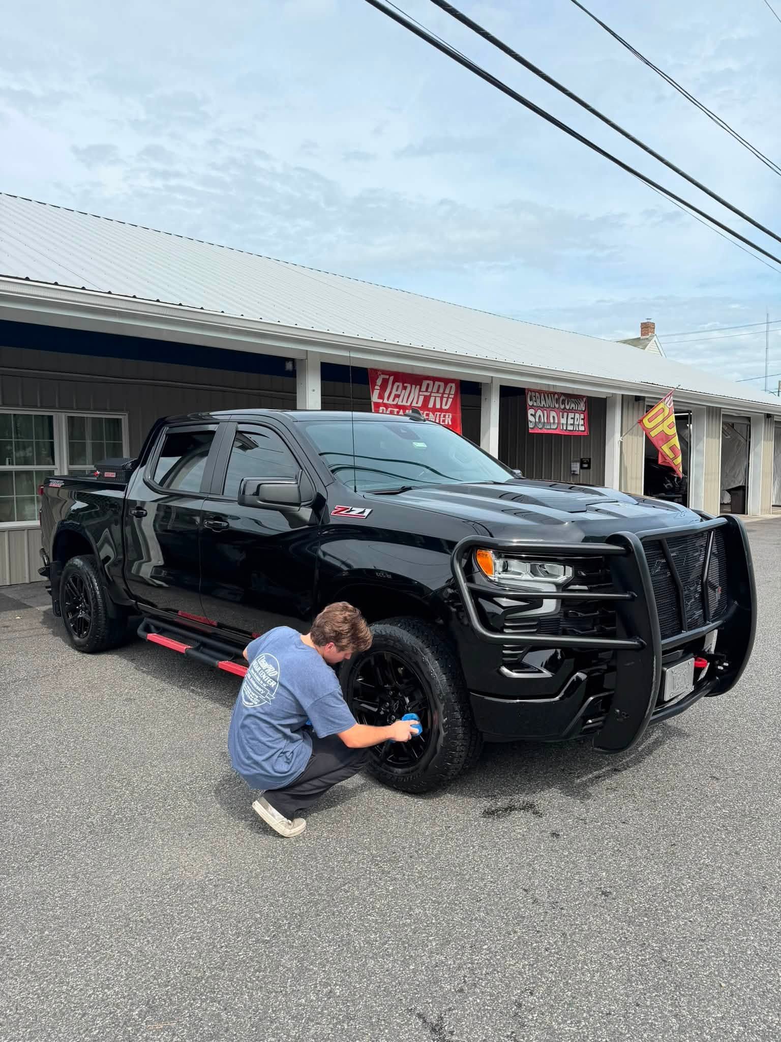 Person crouches beside a black SUV parked outside a storefront, inspecting the front wheel.