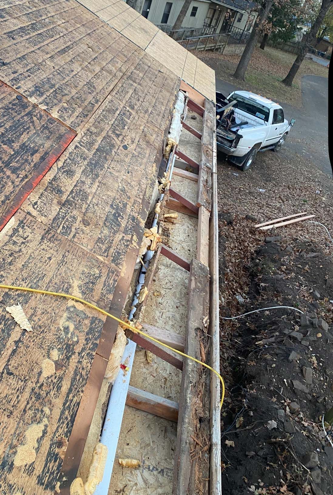 A high-angle view of a roof under renovation, showing exposed rafters, plywood sheathing, and a truck parked below.