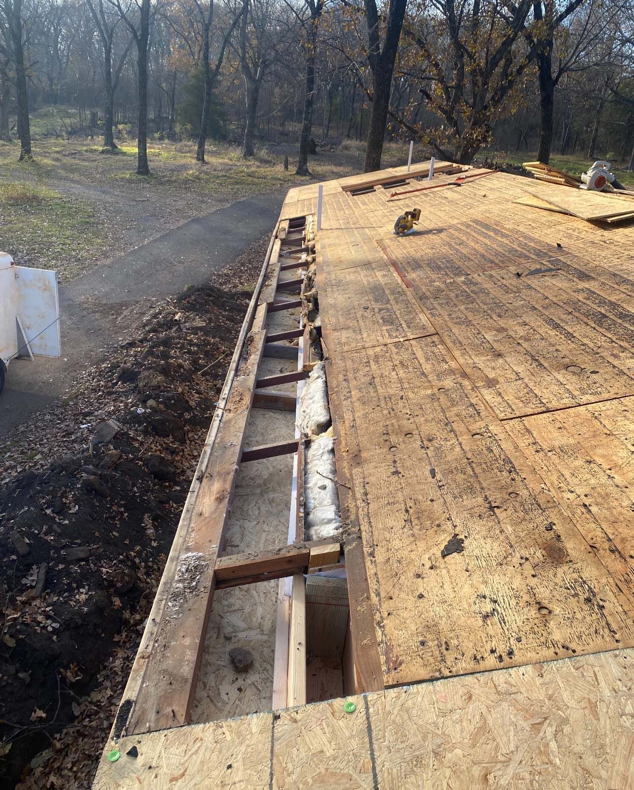 Construction crew repairs an open edge of a wooden roof deck with exposed rafters overlooking a wooded area.