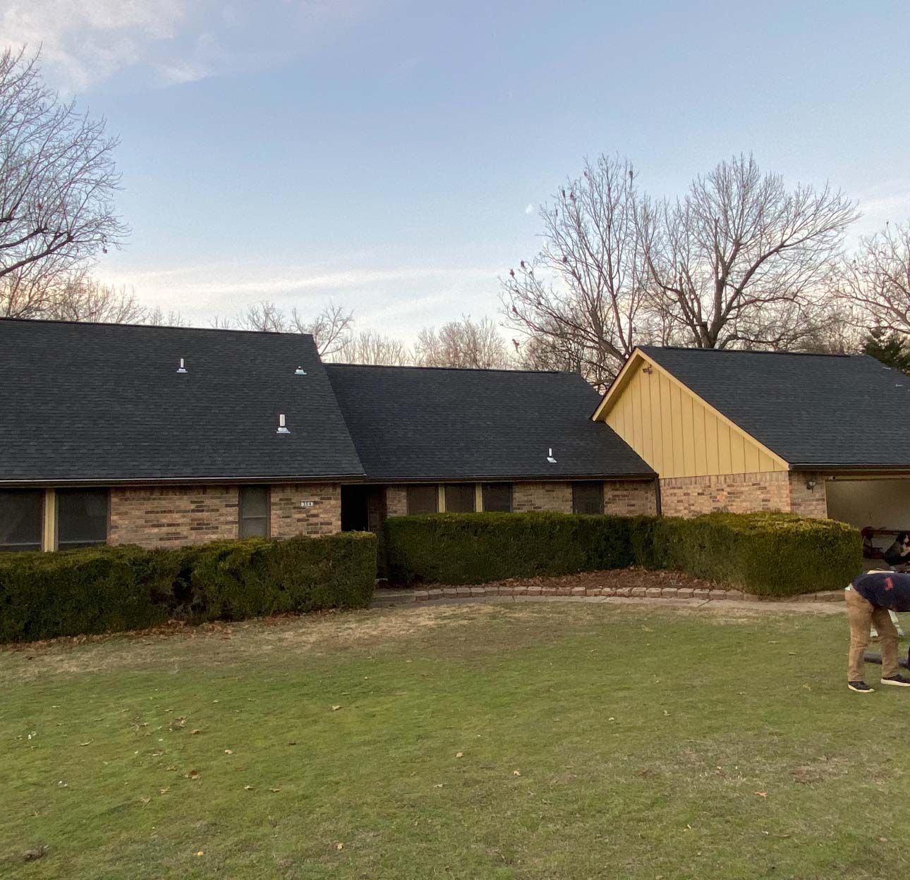 A single-story brick house with a new dark roof and yellow siding, surrounded by hedges and trees on a sunny day.