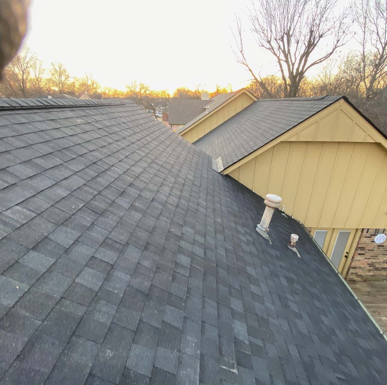 An elevated view of a gray asphalt shingle roof, adjacent to a yellow house exterior with a small vent pipe.
