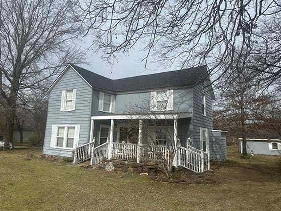 A light blue two-story house with a front porch and a dark roof, set on a grassy lot with leafless trees.