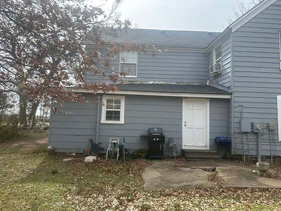 A two-story blue house with a white door, a small window, and a black grill on a concrete patio in a yard with dry leaves.