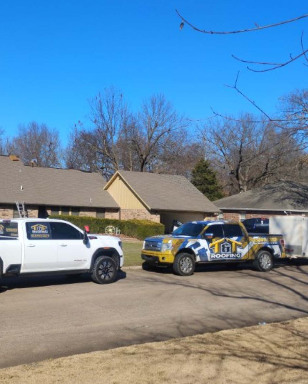 Two work trucks from a roofing company parked in front of a residential house on a clear, sunny day.