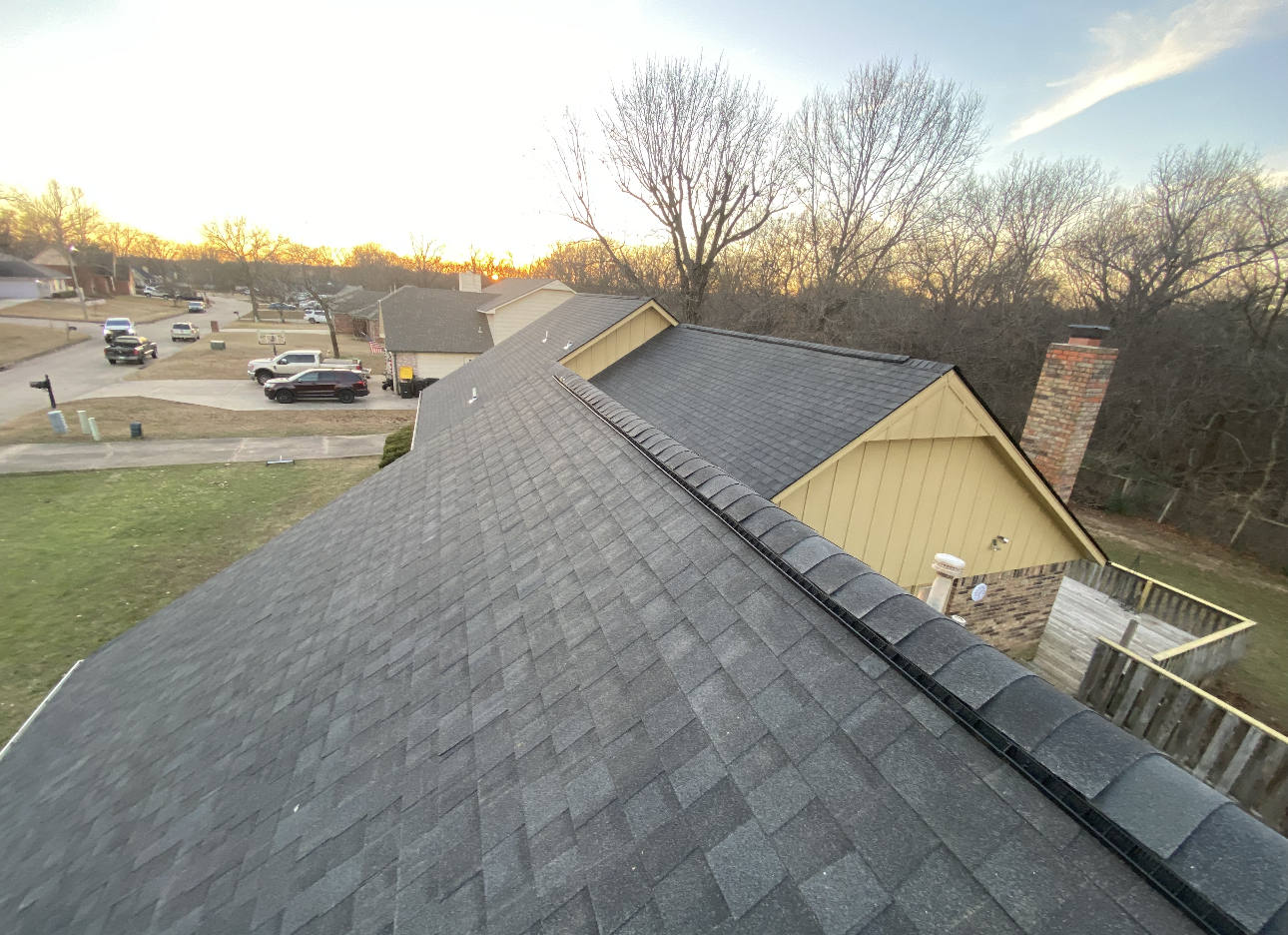 An elevated view of a gray asphalt shingled roof under construction, showing bare yellow wall sheathing and a chimney.