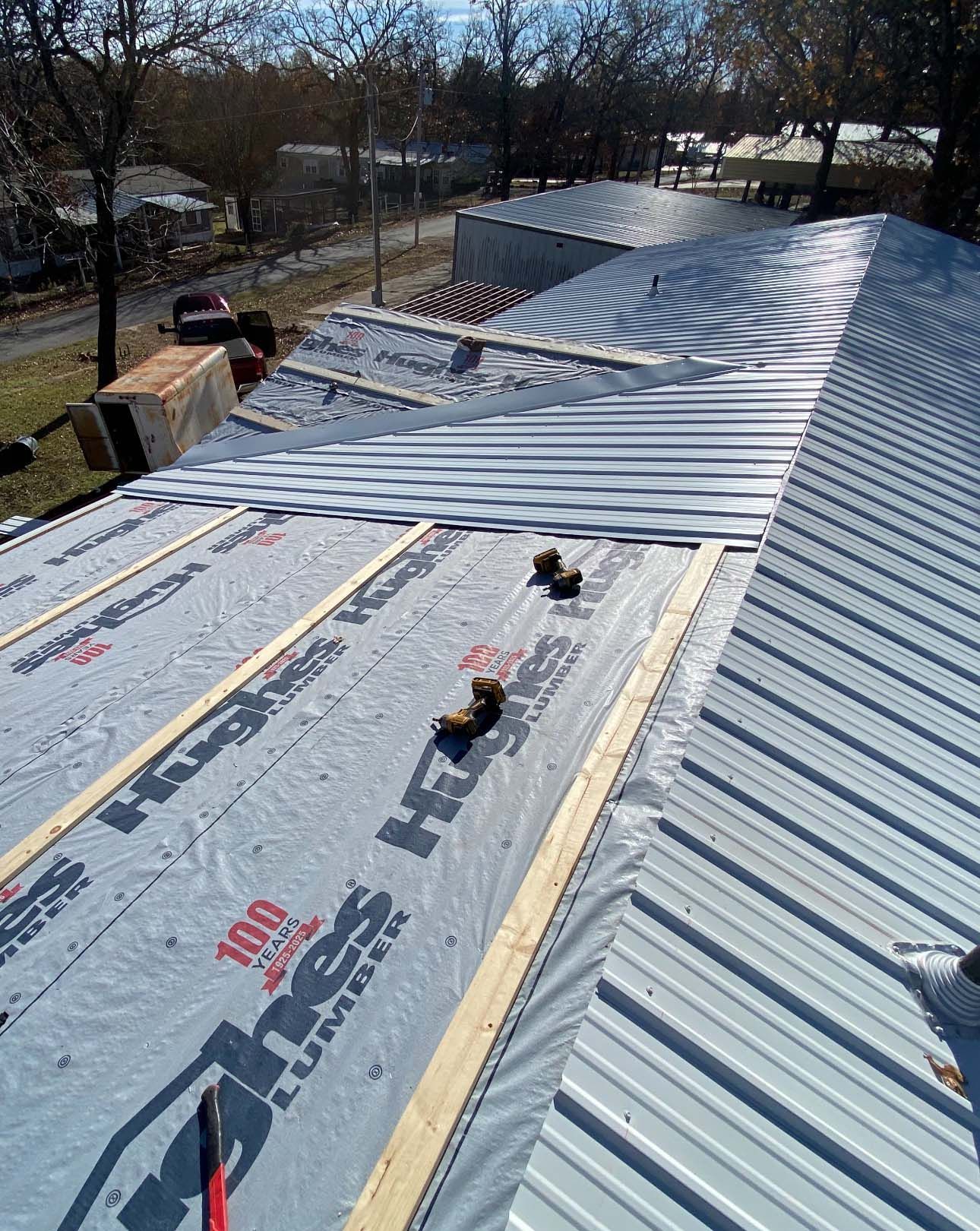A view from a roof showing metal roofing panels being installed over light blue underlayment and wood purlins.
