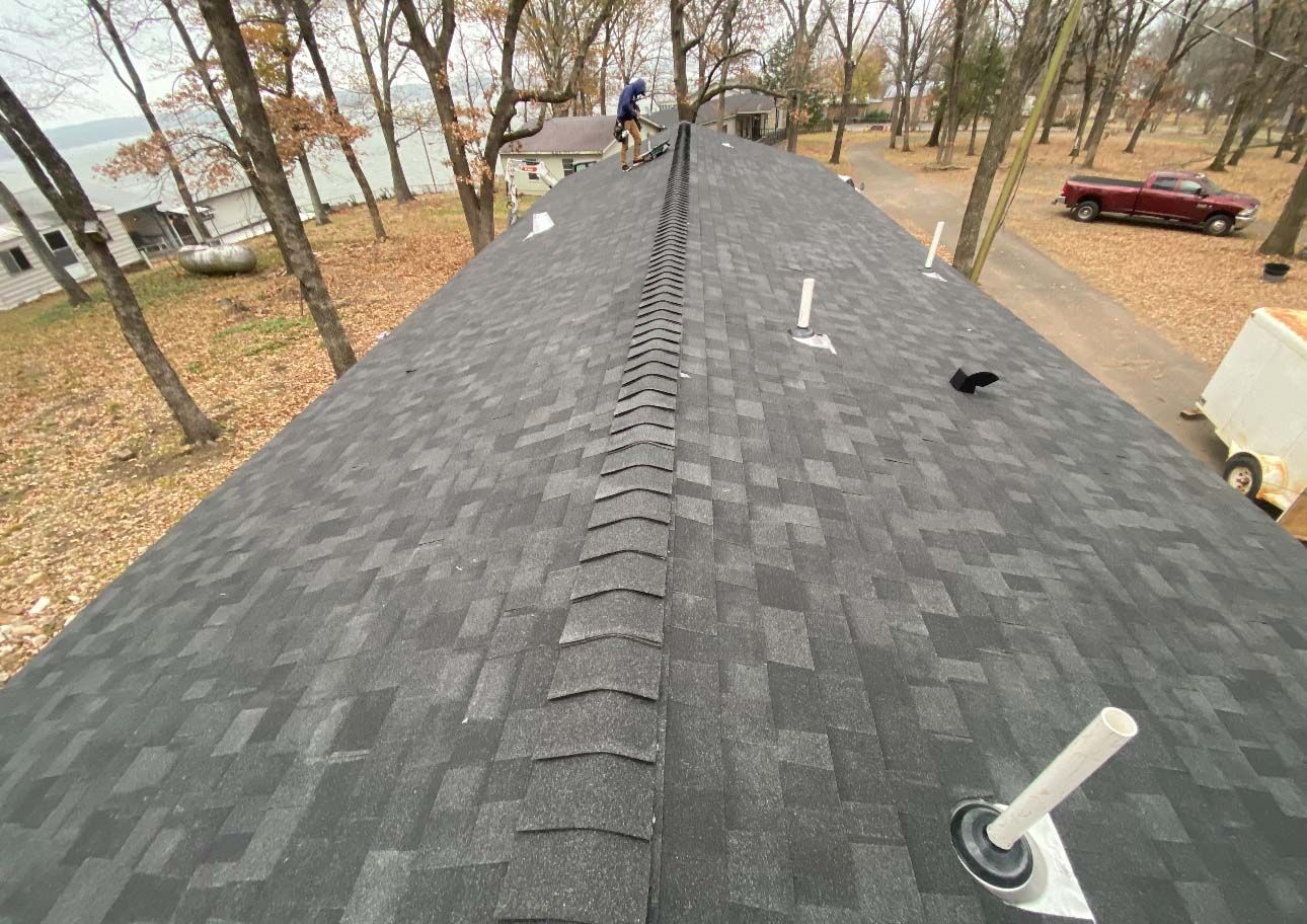 A high-angle view of a newly shingled gray roof with a person standing on the ridge, viewed from above a wooded yard.