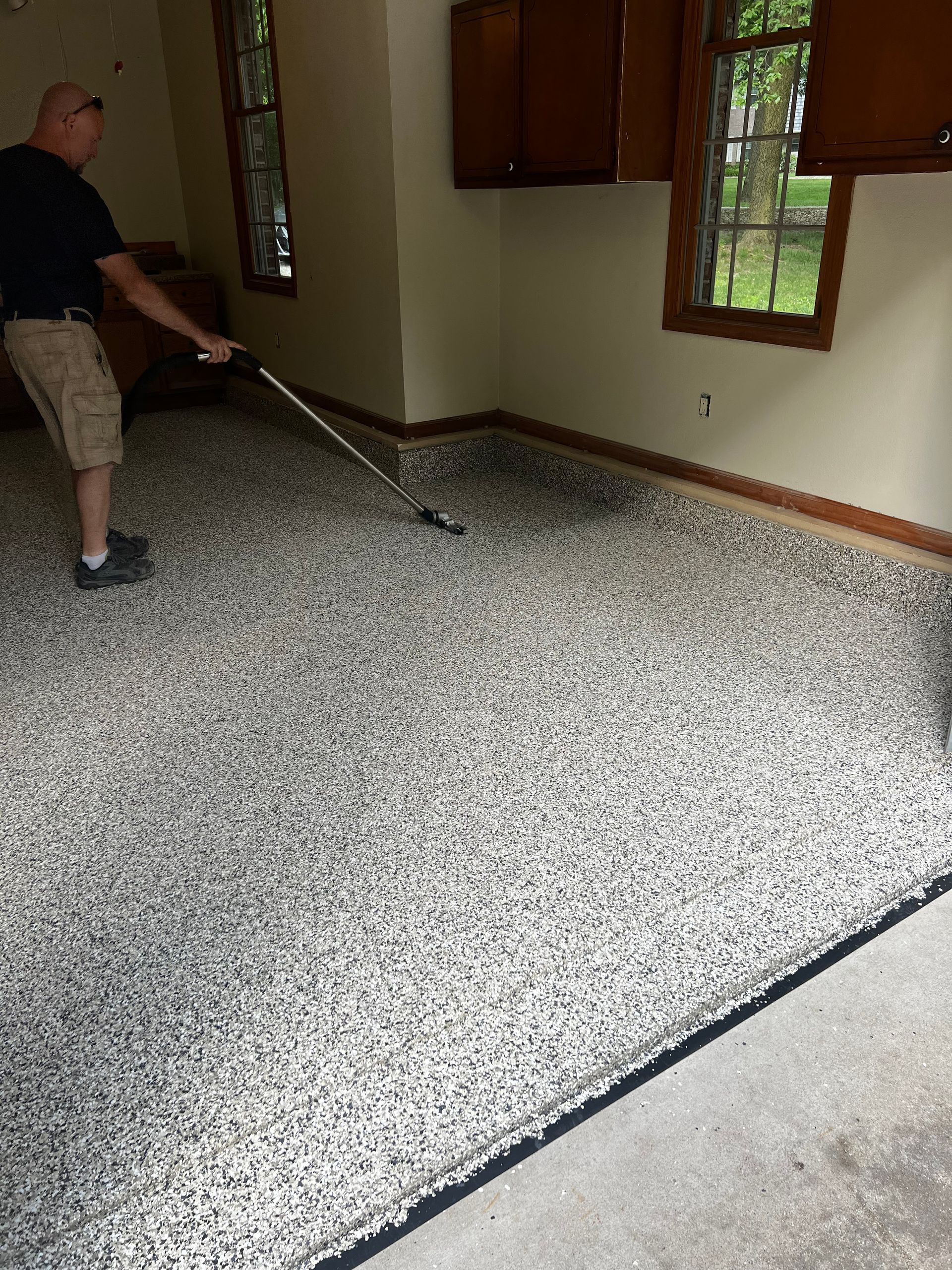 Man raking epoxy-coated garage floor; black, gray, and white speckles, wood cabinets, windows, and light walls.