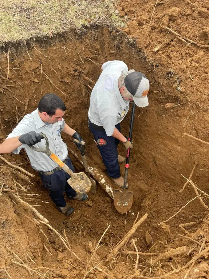 Two workers in a dirt excavation using shovels, standing in a deep trench