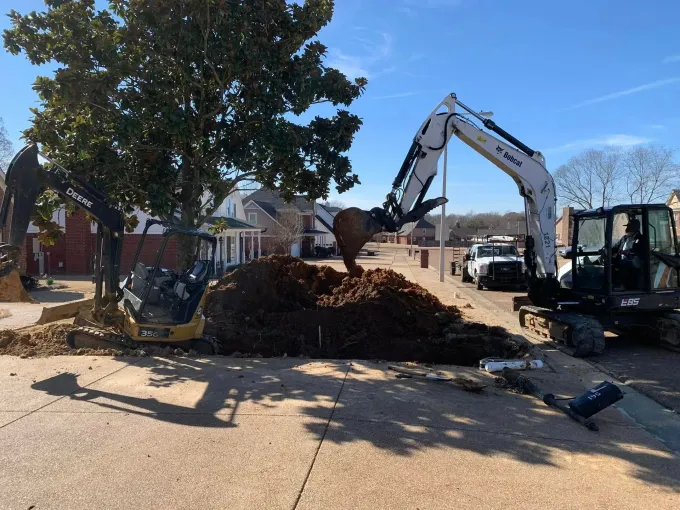 Excavator loading a pile of tree debris on a sunny residential street near houses and parked cars