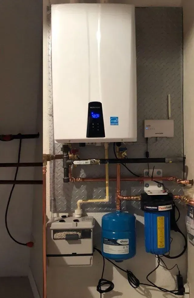 Wall-mounted white water heater with pipes, valves, and blue filtration tanks in a utility room.
