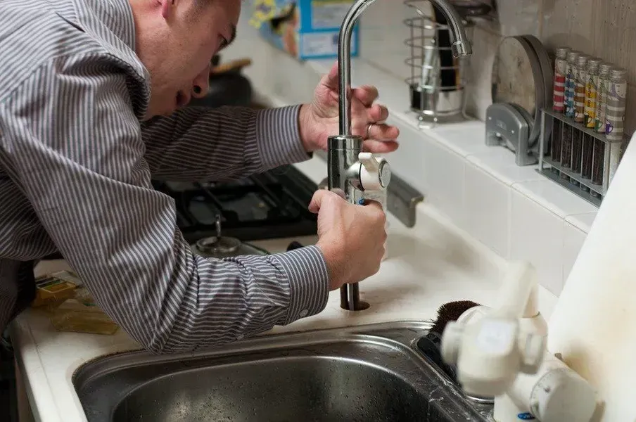 Person repairs a kitchen faucet beside a sink, using tools under the counter.