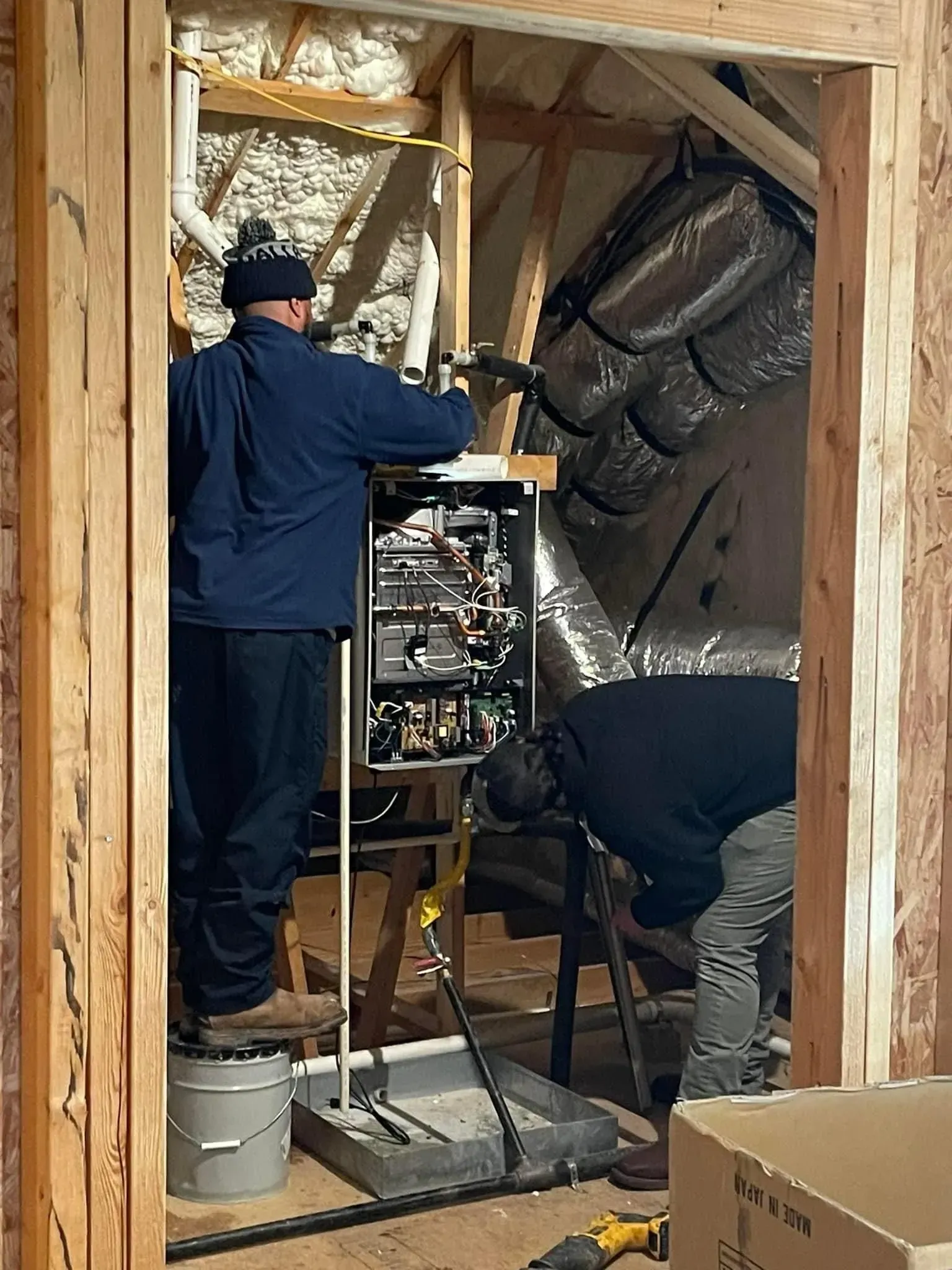 Two workers installing insulation inside a framed wall, one standing and one crouching, with tools and materials nearby
