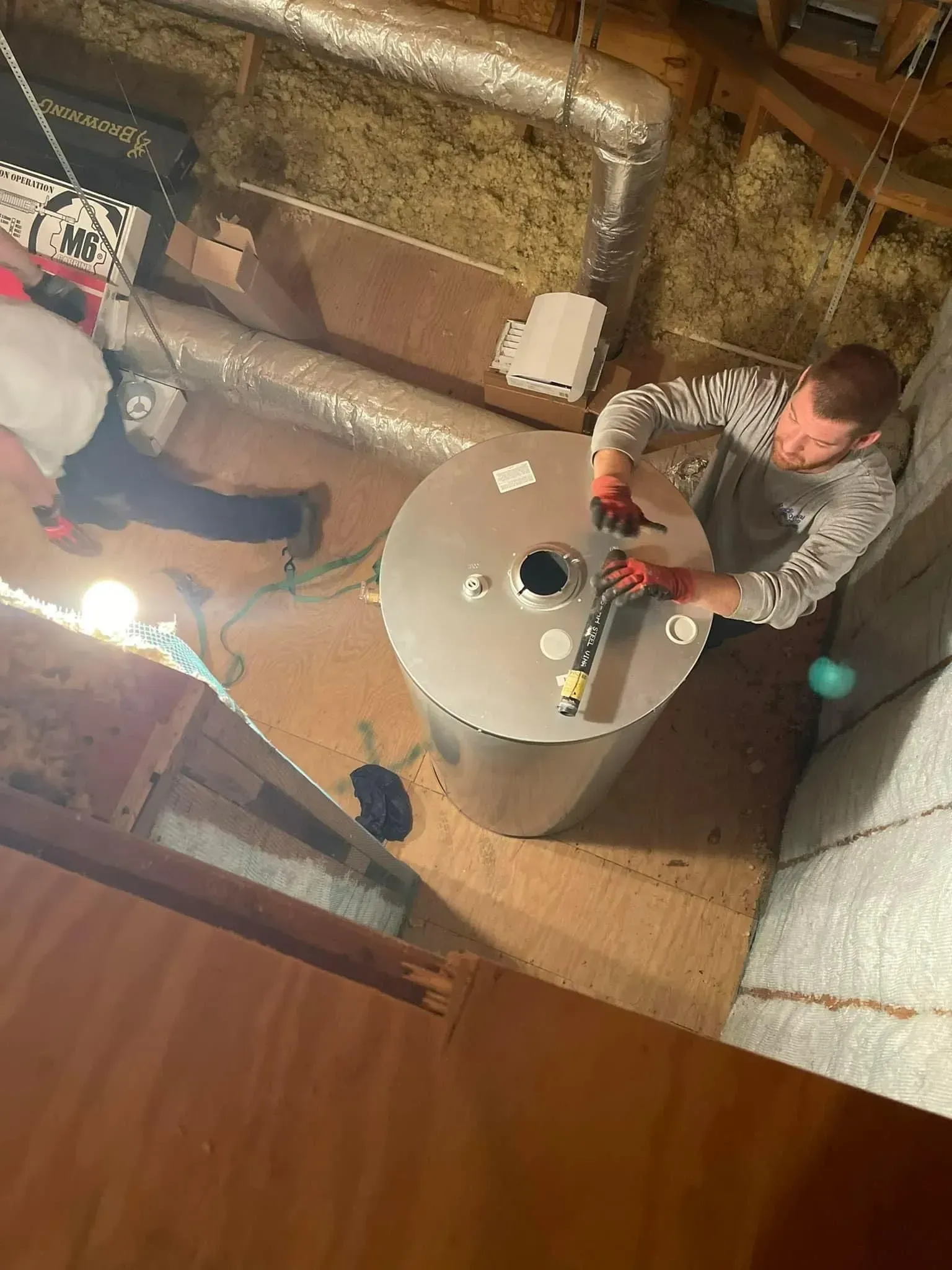 Two people working in a basement near a large white water heater and exposed pipes.
