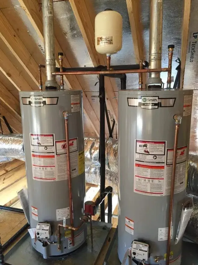 Two gray water heaters in an attic, connected by copper pipes and a small expansion tank.