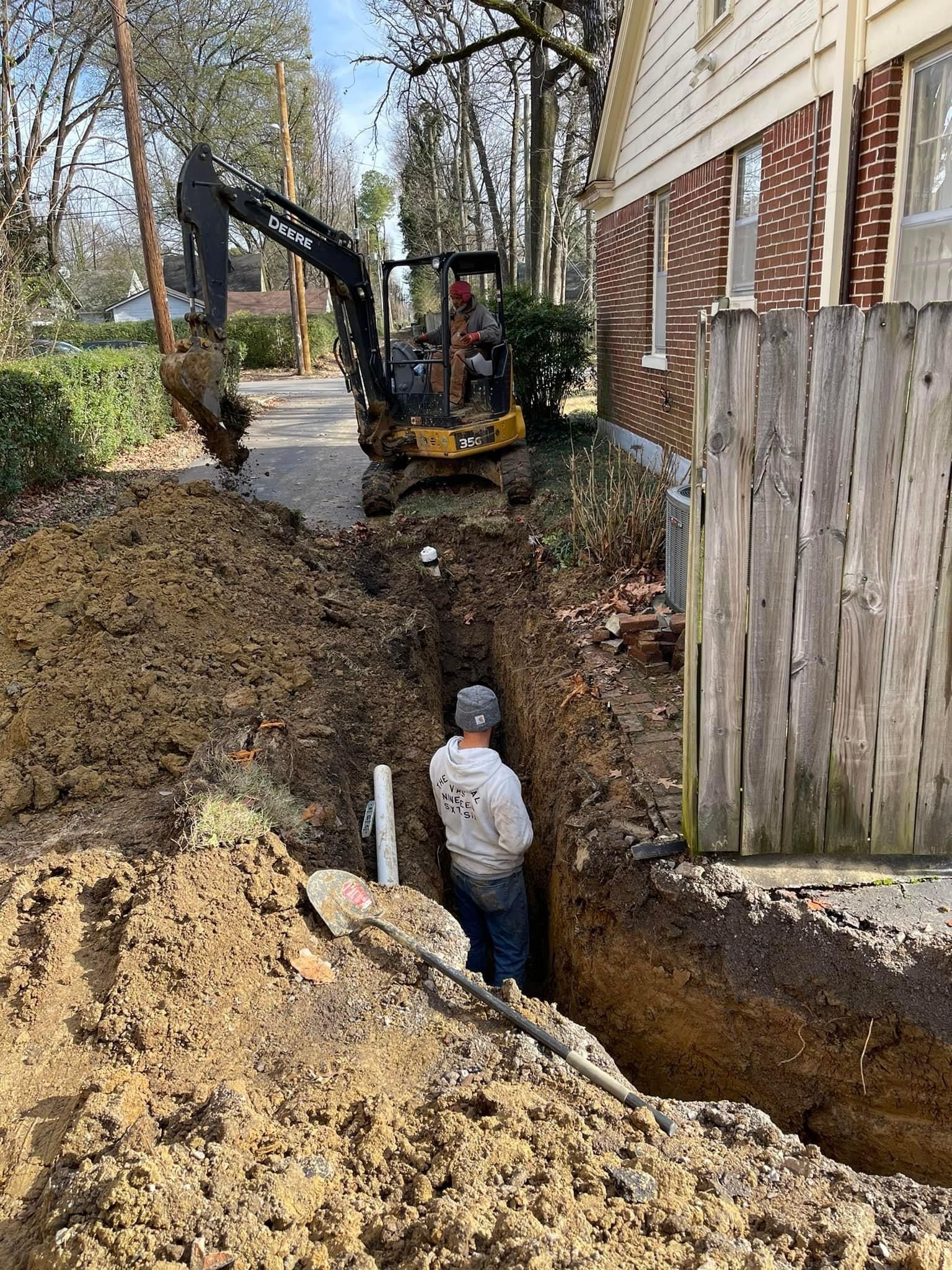 Construction workers digging a trench beside a brick house, with an excavator nearby.