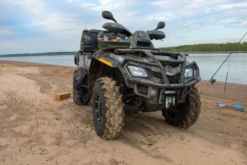 A muddy atv is parked on a sandy beach next to a river.