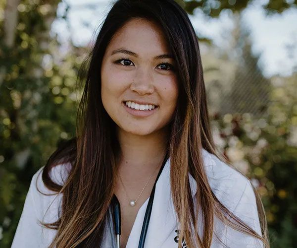 Woman wearing a white coat with stethoscope, smiling outdoors.